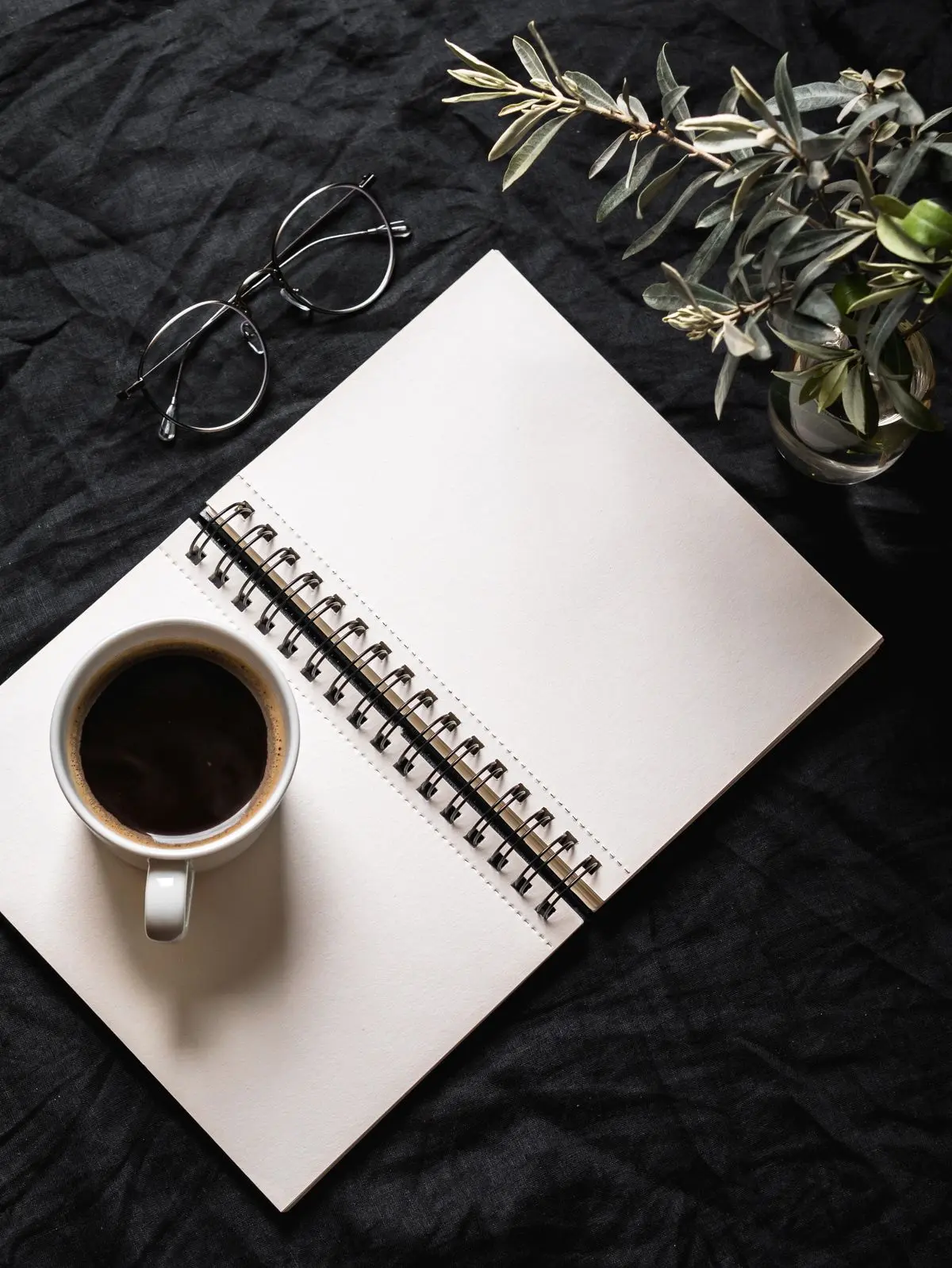 An open notebook, glasses and a cup of coffee beside foliage on dark linen.